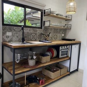 Mueble de cocina con encimera de madera, estante inferior abierto con cestas y utensilios, fregadero negro, estante superior metálico con platos, fondo con azulejos decorativos grises.