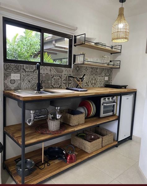 Mueble de cocina con encimera de madera, estante inferior abierto con cestas y utensilios, fregadero negro, estante superior metálico con platos, fondo con azulejos decorativos grises.