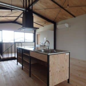 Isla de cocina moderna de madera con encimera de acero inoxidable, estanterías abiertas y grifo. Diseño industrial, situada en un espacio abierto con techo de vigas descubiertas.