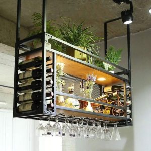 Estante colgante de metal negro con espacio para botellas de vino horizontales y copas invertidas. Incluye lámpara interna, plantas decorativas y compartimento central de malla metálica.