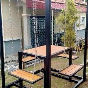 Mesa de madera con bancos colgantes de cadena en estructura metálica negra, ubicada al aire libre en un jardín. Diseño moderno y original para exteriores.