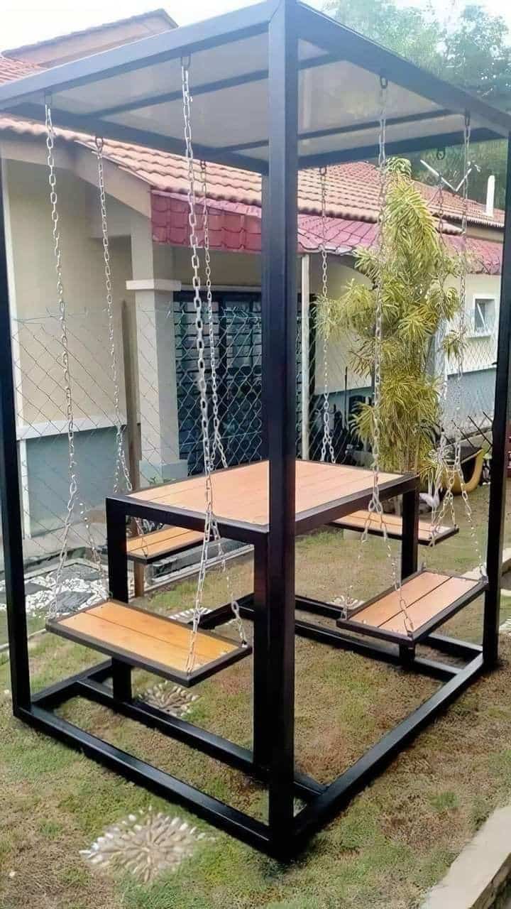 Mesa de madera con bancos colgantes de cadena en estructura metálica negra, ubicada al aire libre en un jardín. Diseño moderno y original para exteriores.