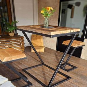 Mesa alta de madera rústica con patas de metal en forma de Z, acompañado de banquetas a juego. Decorada con un jarrón de flores. Ideal para espacios modernos e industriales.