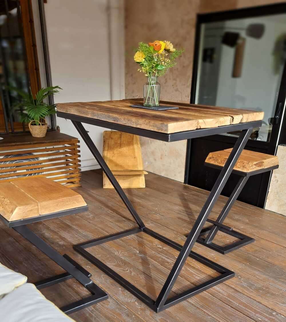 Mesa alta de madera rústica con patas de metal en forma de Z, acompañado de banquetas a juego. Decorada con un jarrón de flores. Ideal para espacios modernos e industriales.