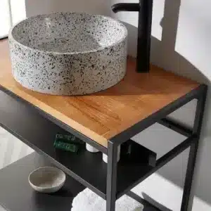 Mueble de baño moderno con estructura metálica negra y tablero de madera clara. Incluye lavabo de granito redondo sobre encimera, grifería negra y estantes para organización.