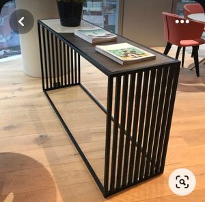 Mesa consola moderna de metal negro con diseño de rejilla, ubicada sobre suelo de madera. Superficie superior lisa y decorada con revistas y una planta.