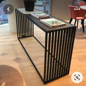 Mesa consola moderna de metal negro con diseño de rejilla, ubicada sobre suelo de madera. Superficie superior lisa y decorada con revistas y una planta.
