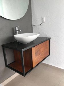 Mueble de baño flotante con estructura negra y puerta de madera. Encimera negra con lavabo blanco de forma ovalada y grifería cromada moderna.