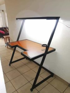 Escritorio moderno de diseño angular en madera y metal negro. Posee estante superior con luz incorporada y espacio para tazas o cables. Ideal para oficinas y estudios.