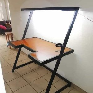 Escritorio moderno de diseño angular en madera y metal negro. Posee estante superior con luz incorporada y espacio para tazas o cables. Ideal para oficinas y estudios.