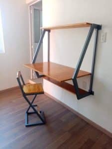 Escritorio de madera suspendido con repisa superior, estructura metálica en triángulo, acompañado de una silla de diseño minimalista, todo sobre suelo de madera oscura.