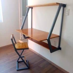 Escritorio de madera suspendido con repisa superior, estructura metálica en triángulo, acompañado de una silla de diseño minimalista, todo sobre suelo de madera oscura.