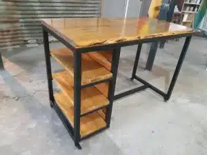 Escritorio industrial de madera y metal, con superficie de madera pulida. Incluye estantería lateral de tres niveles con estantes de madera y estructura metálica negra.