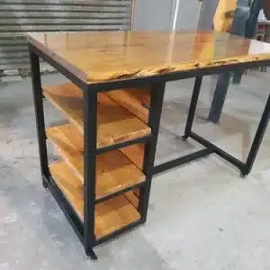 Escritorio industrial de madera y metal, con superficie de madera pulida. Incluye estantería lateral de tres niveles con estantes de madera y estructura metálica negra.