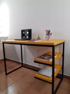 Escritorio moderno de madera clara con estructura metálica negra. Incluye dos estantes laterales y accesorios de oficina sobre la superficie. Ideal para oficinas pequeñas.