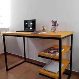 Escritorio moderno de madera clara con estructura metálica negra. Incluye dos estantes laterales y accesorios de oficina sobre la superficie. Ideal para oficinas pequeñas.