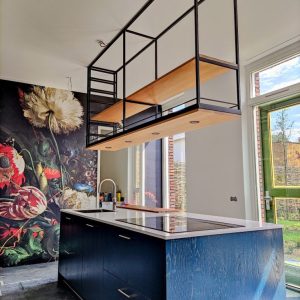 Isla de cocina azul con encimera blanca, integrada con fregadero y vitrocerámica. Encima, estantería suspendida de metal negro con base de madera en techo alto. Diseño moderno y funcional.