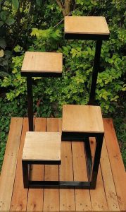 Estante de madera y metal negro con diseño escalonado, ideal para macetas o decoración, colocado sobre una plataforma de madera al aire libre.