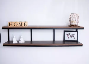 Estante flotante de dos niveles en madera oscura con marco negro. Decorado con letras HOME, figuras blancas, una vela en jaula metálica y un cuadro pequeño.