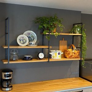 Estante flotante de madera con soportes metálicos negros. Incluye dos niveles decorados con vajilla, plantas, un reloj y una canasta sobre una pared gris de cocina moderna.