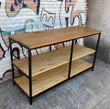 Mesa de trabajo industrial con marco negro de metal y tres estantes de madera clara. Ideal para almacenamiento o exhibición en espacios interiores.