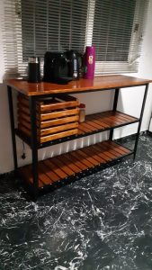 Mesa de madera estilo industrial con estructura metálica negra, estantes inferiores con listones y un cajón de madera en el centro, situada sobre piso marmolado.