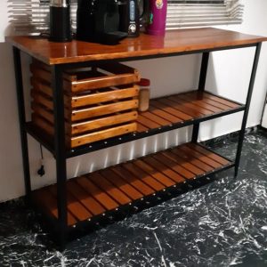 Mesa de madera estilo industrial con estructura metálica negra, estantes inferiores con listones y un cajón de madera en el centro, situada sobre piso marmolado.