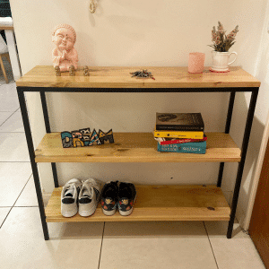 Estantería de madera clara con tres niveles, estructura de metal negro. Decorada con libros, zapatillas y adornos. Ideal para organización y decoración del hogar.