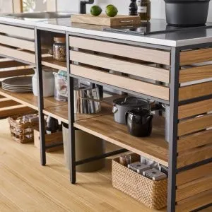 Mueble de cocina moderno de madera y metal, con encimera gris y almacenamiento inferior abierto. Ideal para organizar ollas y utensilios. Diseño funcional y estético en un espacio luminoso.
