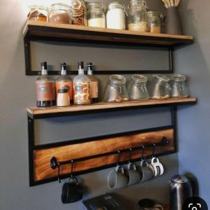 Estantes de madera con soporte metálico en pared gris. Incluyen ganchos para tazas debajo. Adecuados para almacenaje de cocina, combinan estilo rústico e industrial.