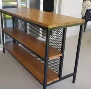 Mesa de diseño industrial con estructura de metal negro y estantes de madera. Cuenta con tres niveles y laterales de rejilla metálica. Ideal para espacios modernos o lofts.