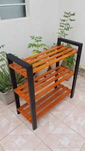 Estante de madera, color naranja con marco negro, de tres niveles, estilo moderno y minimalista. Ideal para plantas o almacenamiento, ubicado en un espacio exterior pavimentado.