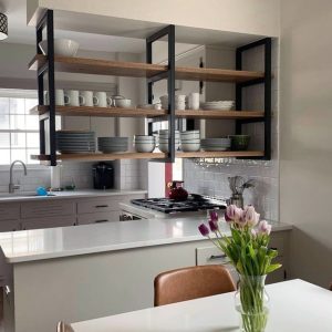 Estantería flotante de madera con soporte metálico negro sobre isla de cocina. Organiza platos, tazas y cuencos, aportando un estilo moderno y funcional al espacio.