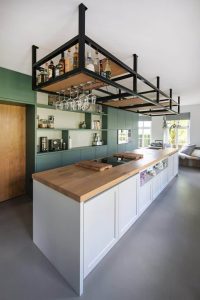 Isla de cocina moderna con encimera de madera clara, gabinetes blancos y rejilla de metal negra suspendida para botellas y copas, combinando funcionalidad y estilo contemporáneo.