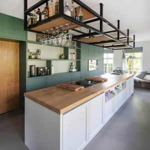 Isla de cocina moderna con encimera de madera clara, gabinetes blancos y rejilla de metal negra suspendida para botellas y copas, combinando funcionalidad y estilo contemporáneo.