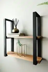 Estante de pared moderno de madera clara con soporte metálico negro. Tiene dos baldas y está decorado con libros y elementos decorativos. Estilo minimalista.