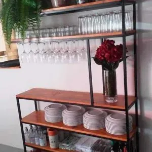Estantería de madera y metal con varias repisas: exhibe cubetas metálicas, copas, vasos, platos, botellas y condimentos. Ideal para organización en cocinas o bares con estilo contemporáneo.