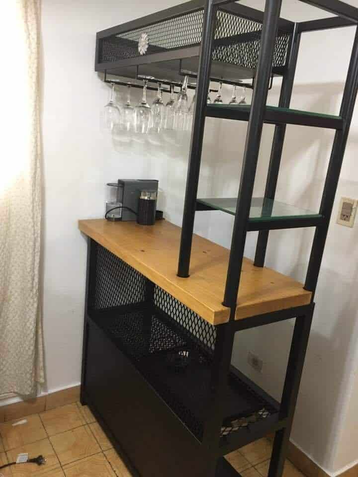 Mueble bar moderno con estructura de metal negro, estantes de vidrio, y superficie de madera. Incluye soporte para copas colgantes y espacio de almacenamiento inferior con rejilla metálica.