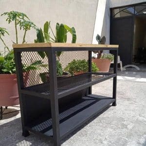 Mesa de estilo industrial con estructura metálica negra, estantes de rejilla y superficie de madera clara. Ideal para uso en exteriores, con plantas de fondo.