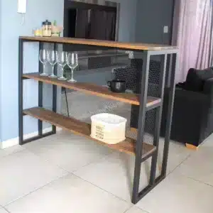 Estante moderno con estructura de metal negro y tres baldas de madera, decorado con copas, jarras y una cesta en ambiente de sala. Perfecto para almacenamiento y decoración.