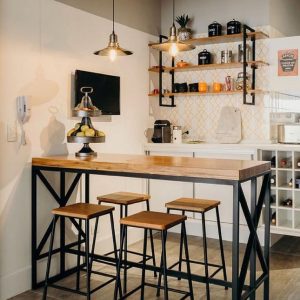 Mesa alta de madera clara con base de metal negro en diseño industrial, acompañada de cuatro taburetes a juego, en un espacio de cocina moderno y minimalista.