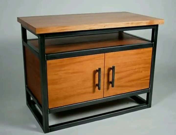 Mueble estilo industrial con estructura metálica negra y paneles de madera marrón. Cuenta con dos puertas y un estante superior abierto. Ideal para almacenamiento en salas modernas.