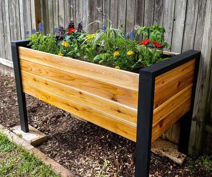 Jardinera elevada de madera con acabado natural y estructura de patas negras, utilizada para plantar flores coloridas. Ideal para jardines de exterior, aportando un diseño moderno y funcional.