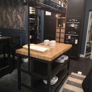 Isla de cocina con encimera de madera clara, estantes metálicos oscuros, soporte superior con plantas y utensilios, diseñada para almacenamiento y preparación culinaria.