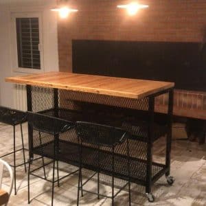 Mesa alta con ruedas, estructura metálica negra y superficie de madera. Incluye cuatro taburetes de metal con diseño de rejilla, ideales para cocina o bar.