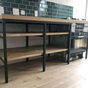 Mesa de cocina con estructura de metal negro, encimera de madera clara y tres estantes abiertos. Ideal para almacenamiento y decoración modernista. Combina funcionalidad y estilo industrial.