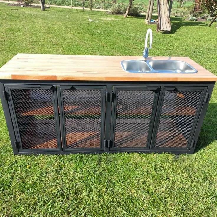 Isla de cocina exterior con encimera de madera, fregadero de acero inoxidable doble y puertas de malla metálica negra. Ideal para espacios al aire libre.