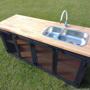 Gabinete de cocina para exterior con fregadero doble de acero inoxidable, encimera de madera clara y puertas de rejilla metálica negra, ideal para jardines o terrazas.