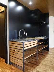 Cocina moderna con fregadero de acero inoxidable, encimeras de madera, estructura metálica negra y estantes abiertos. Diseño minimalista y funcional en espacio compacto.