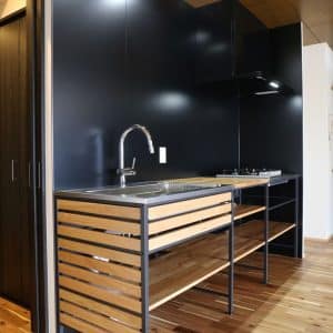 Cocina moderna con fregadero de acero inoxidable, encimeras de madera, estructura metálica negra y estantes abiertos. Diseño minimalista y funcional en espacio compacto.